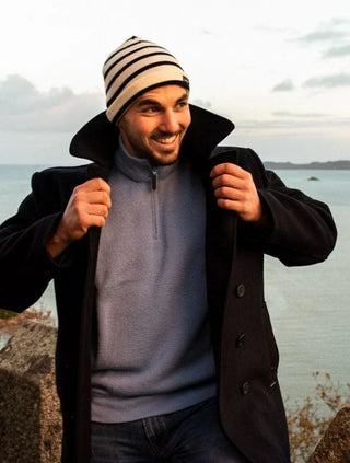 Man wearing a striped beanie, navy coat, and blue pullover by the water