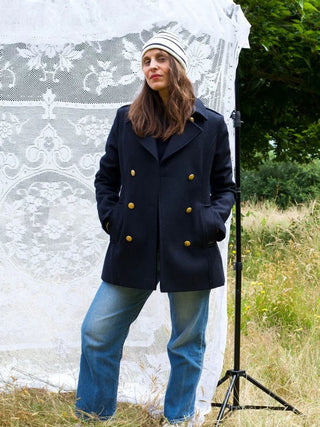 Woman in navy peacoat, striped beanie, and jeans posing outdoors in front of lace backdrop
