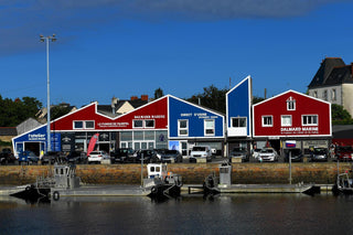 Row of colorful marine-themed shops with red and blue facades by the waterfront and parked cars