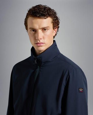 Man wearing navy blue nylon zip-up jacket with high collar and small chest logo patch
