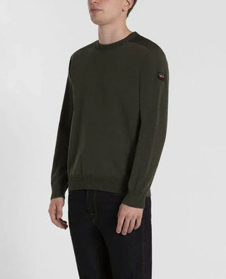 Man wearing olive green crew neck sweater with shoulder patches and jeans, standing indoors