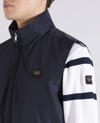 Navy nylon sleeveless vest with zip front and logo patch, worn over striped white sweater