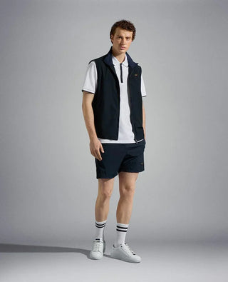 Man wearing a navy nylon vest, white polo shirt, navy shorts, and white sneakers