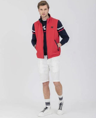 Man wearing a red sleeveless nylon vest, navy sweater, white shorts, and sneakers