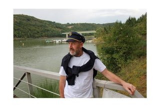 Man in navy captain hat and casual outfit by a river with forested hills in the background